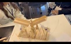 A photo showing a popsicle stick bridge with a flat, rectangular base. There is a syringe on each side of the base that is being held together with popsicle sticks attached to base and syringe at an angle. Each syringe is attached to a plastic tube that had an additional syringe on the other end to control movement through the tube. The parts of the syringe that get push and pulled are directed toward the top portion of the bridge. The top portion of the bridge has 2 leaf-like parts that lift when the syringes are pushed on each side of the top platform, and when the syringes are pulled, the platform lowers.