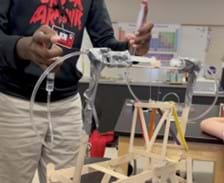 A photo showing a student pushing 2 syringes, one in each hand, attached to a plastic tube with a horizontal syringe on each side. The horizontal syringes are attached using duct tape to a thin skewer that has a small cube on it. Attached to the cube is a thin string holding a small platform made from small popsicle sticks. The skewer holding all of this is attached to a rectangular base made from popsicle sticks, with 2 longer popsicle stick towers (held together by duct tape) standing above the bottom base. As the student pushed one syringe, the syringe at the other end of the plastic tube extends pushing the block the other direction, and when the student pushes the opposite syringe, the same thing happens but in the other direction.