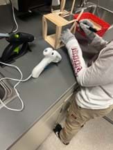 A photo showing a student measuring popsicle sticks glued together with the goal of attaching it to the base of their bridge. The work area is clean, with two hot glue guns and a supply basket visible. The student is also using protective gloves while constructing their bridge.