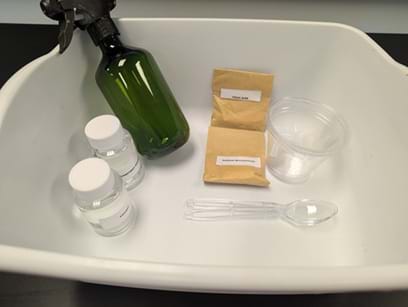 A photo of a white plastic bin containing materials for a hands-on activity. Inside the bin are a green spray bottle, two small clear bottles with white lids, two sealed brown paper packets with labels, a clear plastic cup, a plastic spoon, and two plastic pipettes. The items are arranged loosely inside the bin on a dark tabletop.