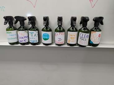 A photo of eight dark spray bottles with black trigger sprayers lined up on a whiteboard ledge. Each bottle has a colorful hand-drawn label identifying different cleaning solutions, including names such as “Awesome Dark,” “RA,” “Super Soaker,” “Clean Max,” and “Cool.” The labels use different colors and drawings, indicating the bottles contain different mixtures. The whiteboard behind them has faint marker drawings.