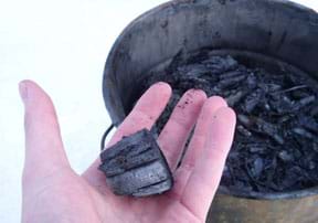 A photograph shows a hand holding a lump of black charcoal pulled from a pot of the same material sitting nearby.