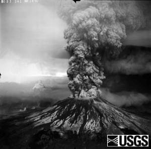 A black and white photo shows a large and thick column of gas and ash erupting out of a volcano’s peak, straight up into the air above the mountain.