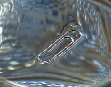 A photograph shows a steel paper clip floating on a water surface. The clip distorts the water surface slightly, but does not sink.