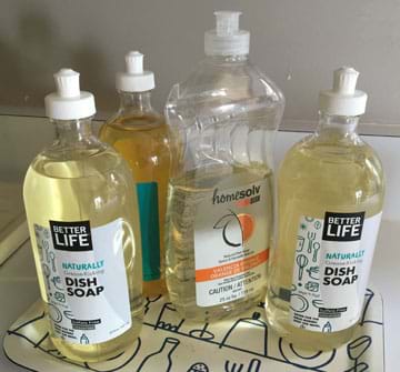 A photograph shows four plastic squeeze-top capped clear plastic bottles containing three different brands of liquid hand dish soap sitting on a tray on a kitchen counter.