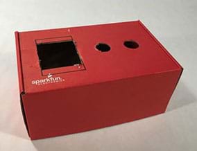 A photograph shows a red-painted cardboard box with three holes cut in its top side—a square hole and two smaller round holes.
