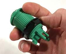 A photograph shows a hand holding a threaded green and black plastic device (same as in Figure 4; plastic arcade button) tilted slightly to show the thumb moving the black plastic nut (looks like a ring) to unscrew it from the button.