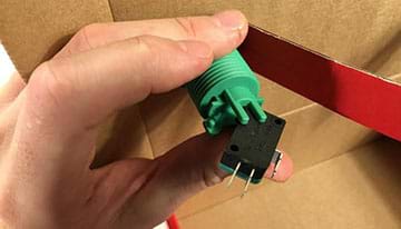 A photograph shows a hand inside the shoebox holding the green arcade button base and reconnecting the black plastic switch to it.