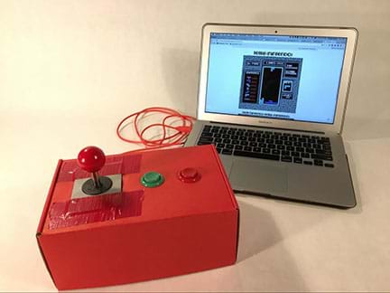 A photograph shows a red-painted shoebox with a red-ball joystick and two big, round plastic arcade push buttons (green, red) mounted on its top surface. A red wire connects the box to a nearby laptop that has a video game interface on its monitor.