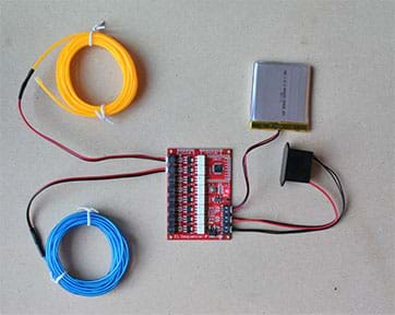 A photograph shows coiled lengths of blue and yellow EL wires each connected to a red EL sequencer circuit board. Also connected to the board is a battery that powers the sequencer.