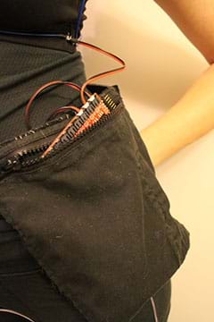 A close-up photograph shows a jacket pocket sewn on a belt being worn on a girl’s hip. Inside the pocket pouch is the EL sequencer (red circuit board) with its wires protruding out of the pocket.