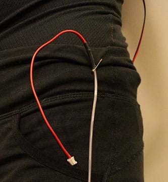 A photograph shows a safety pin holding a strand of red EL wire to the clothing on a woman’s back hip. 