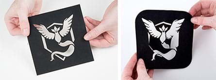 Two photographs show hands holding the same Pokémon Articuno character stencil cut from square black iron-on fabric patches. The stencil cutout looks like an abstract flying bird and fluttering ribbon in front of a diamond-shaped shield. The stencil on the right shows some of the thin and delicate shield shape cutouts ripped.