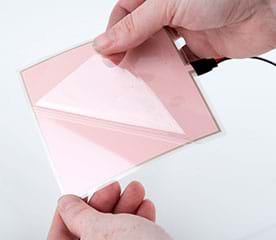 A photograph shows a hand holding a shiny 10 x 10-cm square EL panel while another hand peels away a thin, clear plastic cover sheet from the EL panel.