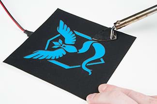 A photograph shows a hand using the point of a mini iron to warm the cut-out fabric patch stencil placed over an EL panel so as to activate the patch adhesive. Under the cut-out black stencil (and on the EL panel) is a layer of blue lighting gel, which reveals a Pokémon Articuno species graphic that looks like a stylized blue bird with outstretched wings and a ribbon tail in front of a diamond-shaped shield.