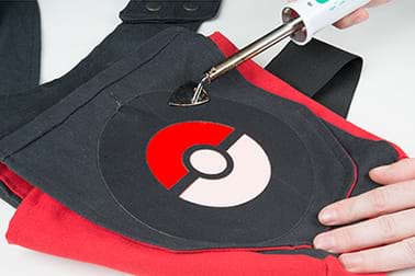 A photograph shows a mini iron being used to warm the fabric adhesive/adhere a Poke ball patch onto the flap of a messenger bag.