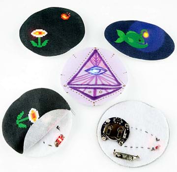 A photograph shows three completed light-up pins (flower and sun, fish, pyramid with eye). The flower pin is also flipped over to show its battery pack on the back that provides an electricity source, and a pin to attach it to clothing. Another photo of the same pin shows its front fabric layer pulled back to reveal its middle layer with a sewn-in inner circuit and LED light on felt.