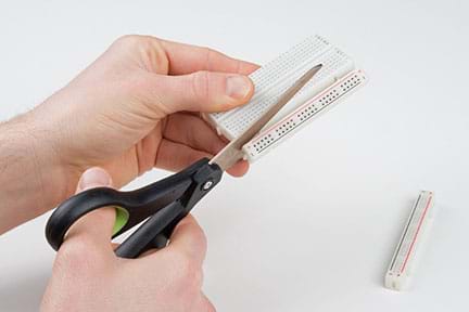 A photograph shows two hands using a pair of scissors to cut off the striped ends of a breadboard.
