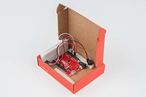 A photograph shows a lidded cardboard box containing the breadboard with the speaker, the RedBoard, and the breadboard with the potentiometer, all connected with wires.
