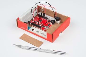 A photograph shows a cardboard box containing a RedBoard, breadboard with speaker, a breadboard with potentiometer, and notches on the front and right box sides to permit keyboard and power cord access.