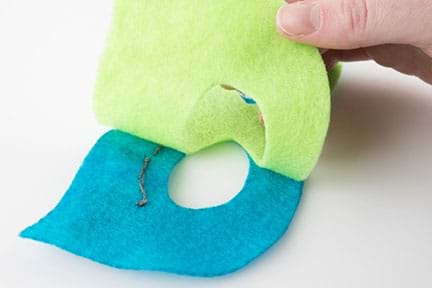 A photograph shows a hand placing a larger green felt eye mask on the back of a slightly smaller blue felt eye mask that has stitching on it.