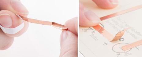 Two photographs. One hand holds the end of a strip of copper tape while another hand’s fingers bend back about a half-inch of the end of the tape so it sticks to itself. This is done at a location on a paper circuit so that the flap of copper tape is positioned over a circle on the template diagram, which represents the coil cell battery location.