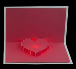 A photograph shows a hand opening a red and white pop-up and light-up valentine card that says “I <heart> you.”