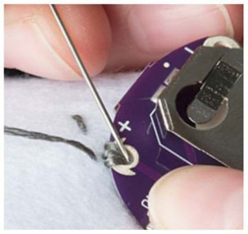 A close-up photograph shows fingers poking a threaded needle into a grommet hole (positive pin) of a battery holder, making a few loops of conductive thread to sew it to the felt backing.