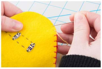A photograph shows a hand holding a felt monster cutout with its sewn circuitry visible and another hand using a needle with colored embroidery thread to stitch up the outer seam, which joins the two sides together in order to close it up for stuffing.