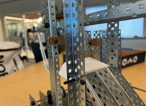 A photo showing a silver steel machine built with VEX parts that has a combination of wheels, gears, levers, and inclined plane. Two parts have been replaced with brown cardboard, and one replaced with white cardstock.