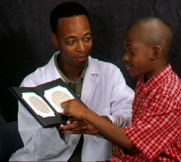 A photograph shows a man in a white coat sitting next to a young boy in a dark room. The doctor holds two color plates and waits for the child to tell him what he sees.