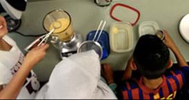 A photograph from above a classroom counter with a blender, mesh sieve and many containers shows three students crowding around the algal cell edible model setup.