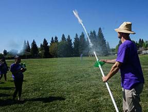 A photograph shows a person in a grassy schoolyard holding a long white PVC pipe, which is a water bottle rocket launcher. The person launches a rocket into the sky while two students observe in the background.