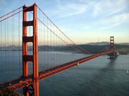 A photograph shows a two-mile long reddish orange cable bridge spanning a wide water channel. It has two tall towers and roadway supported by a metal truss made of triangular shapes.