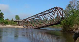 A photograph shows a collapsed steel truss bridge with one end sinking into the middle of a small river. A photograph shows a collapsed steel truss bridge with one end sinking into the middle of a small river.