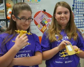 Two young people stand in a classroom, each smiling and holding in their hands four-wheeled vehicles made of dry pasta. 