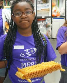 A photograph shows a young person holding in one hand a small, two-axle vehicle made of various types of dry pasta. It looks like a long raft of lasagna noodles with spaghetti axles and rotelle wheels.