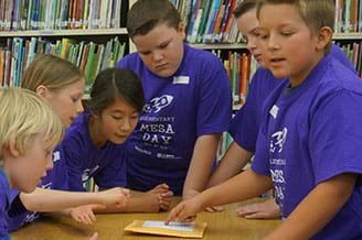 A photograph shows six students around a table, all focused on a drawing as they brainstorm their obstacle course strategy.