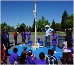 A photograph shows at ~15 young students standing and sitting around a square blue tarp on which an egg dropper rig is placed. The rig is a 10-foot-tall wooden structure with an arm and pulley. Falling from the rig, a white egg is seen midair above a student-designed catcher on the blue tarp.