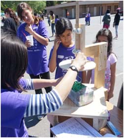A photograph shows young students watching a teacher measure an egg catcher (looks like a box filled with shredded paper) by using a homemade wooden tool that consists of a 25-cm wood circle and a sliding ruler. A student team hopes its egg catcher is not too big!