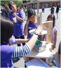 A photograph shows young students watching a teacher measure an egg catcher (looks like a box filled with shredded paper) by using a homemade wooden tool that consists of a 25-cm wood circle and a sliding ruler. A student team hopes its egg catcher is not too big! A photograph shows young students watching a teacher measure an egg catcher (looks like a box filled with shredded paper) by using a homemade wooden tool that consists of a 25-cm wood circle and a sliding ruler. A student team hopes its egg catcher is not too big!