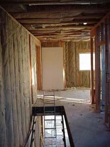 A photograph shows the inside upstairs corner of a wood-framed house with the long spaces between the wall 2 x 4s and ceiling joists filled with paper-backed fiberglass insulation (before drywall installation).