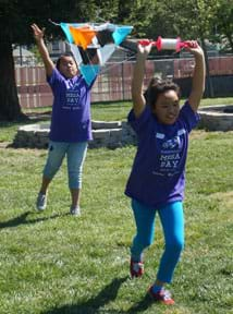 A photograph shows two young girls in a grassy playground attempting to launch a kite. One girl tosses a multi-celled tetrahedron kite high into the air as her partner runs ahead of her holding above her head a spool of string connected to the kite.