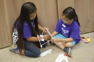 A photograph shows two young girls sitting on the floor constructing a sail car with a square frame base made of drinking straws, with wheels at the corners. On top of the frame, a three-sided pyramid made of straws supports a triangular sail.