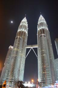 A photograph at night shows two tall, pointed twin skyscrapers connected by a mid-way pedestrian bridge, with the dark sky and moon in the background.