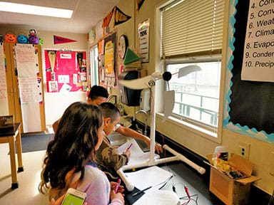 A photograph shows three youngsters at a long counter where a model wind turbine is set up. The turbine has four cardboard blades that re spinning. One boy is writing on his worksheet.