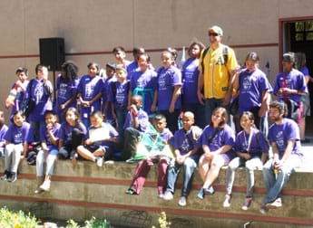 A photograph shows a group of 20+ students in matching purple T-shirts posed in a group outside a school on an award stage; they are competition participants from the winning school.