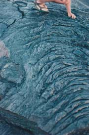 Photo shows a person standing on a wide swath of black rock that is textured with ripples and ridges.