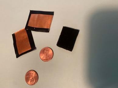 Two one-inch square pieces of copper flashing with electrical tape around 3 sides, two pennies, and one piece of black conductive foam approximately the same size as the copper.
