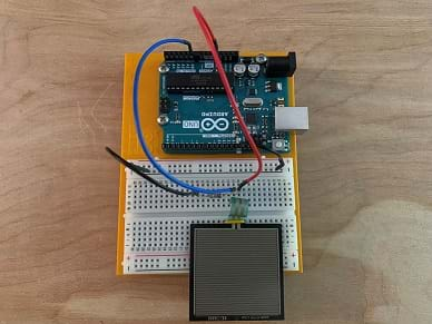 A square force-sensitive resistor is connected via a voltage divider circuit with a 10 k resistor built on a solderless breadboard and connected to an Arduino microcontroller.